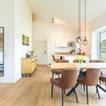 Essbereich mit Holztisch und braunen Stühlen. Küche und Wohnbereich mit Holzboden und großen Fenstern.