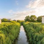 A canal flows between green banks and houses under a blue sky.