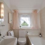 Badezimmer mit Badewanne, Toilette und Waschbecken. Wandfliesen mit Muster und Fenster mit rosa Vorhängen.
