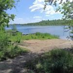 A beach area by the lake with trees and a pavilion in the background.