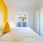 Schlafzimmer mit Bett, gelber Wand und Bild. Tür führt nach draußen.