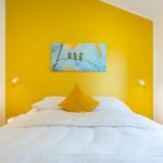 Schlafzimmer mit gelber Wand, Bett mit weißer Bettwäsche und gelbem Kissen, Wandbild mit Vögeln.
