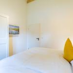 Schlafzimmer mit Bett, gelber Kissen und Fernseher an der Wand.