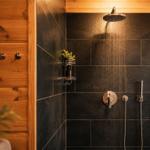 Dusche mit schwarzen Fliesen und Holzwand. Duschbrause und Handbrause sind sichtbar.