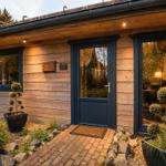 Holzhaus mit Eingangstür und Glasfront. Beleuchtete Außenwand und Pflanzen im Vorgarten.