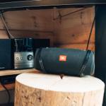Ein JBL-Lautsprecher steht auf einem Holzstumpf neben einem Panasonic-Receiver.