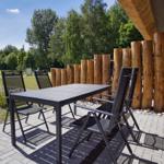 Terrasse mit Tisch und Stühlen, Holzpfosten und Ziegelwand. Grünfläche und Bäume im Hintergrund.
