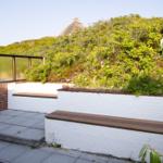 Terrasse mit Sitzbänken, weißen Mauern und grüner Vegetation. Im Hintergrund ist ein Dach zu sehen.