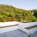 Terrasse mit weißen Mauern und Holzbänken, umgeben von grüner Vegetation und einem Steinweg.