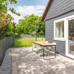Terrace with table and chairs beside gray house. Backyard with grass and fence.