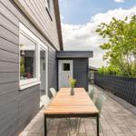 Outdoor area with table and chairs in front of gray wooden house. Greenery and fence in background.