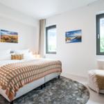 Schlafzimmer mit Bett, Stuhl und Fenstern. Wandbilder zeigen Strandlandschaften.