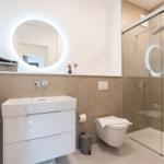 Badezimmer mit Dusche, Waschbecken und Toilette. Rundes Spiegellicht und beige Fliesen.