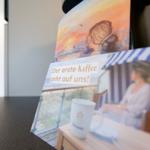 Kaffeebecher auf Tisch neben Bildern mit Meerblick und Sonnenuntergang.