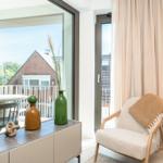 Ein Raum mit Sessel, Tisch und Blick auf Balkon. Vorhänge und Vasen sind sichtbar.
