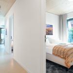 Schlafzimmer mit Bett, Fenster und Decke aus Holz. Durchgang zu Wohnbereich mit Tisch und Stühlen.