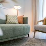 Ein Wohnbereich mit Sofa und Sessel, Bezug in Grün und Beige, Kissen mit Muster, Wandlampe und Fenster mit Vorhängen.