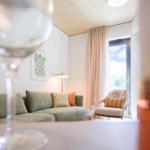 Wohnzimmer mit Sofa, Sessel und Fenster. Vordergrund: Weinflasche und Glas.