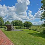 Roter Backsteinbau mit Terrasse, Grasfläche und Blick auf Felder unter blauem Himmel mit Wolken.