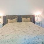 Schlafzimmer mit Bett, Fenstern und Gardinen. Lampe und Schrank sind sichtbar.