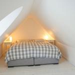Hölzerner Flur mit weißer Brüstung und schrägen Wänden. Durchgang zu einem Schlafzimmer mit Bett.