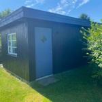 Wooden house with blue door and window, surrounded by grass and shrubs under blue sky.