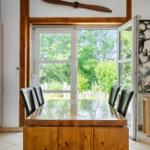 Dining room with wooden table, chairs, and view of green landscape through glass doors.