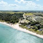 Beach with white sand and clear water, surrounded by houses and green areas.
