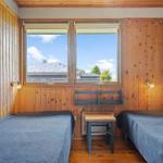 Two beds with blue bedspreads, wooden walls, and windows with a view of roofs and trees.