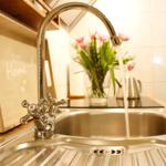 Küchenarmatur mit laufendem Wasser, Holzbrett und Blumen im Hintergrund.