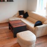 Ein Wohnzimmer mit cremefarbenem Sofa, Holztisch und Fußhocker. Wandregal mit Dekoration.