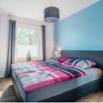 Schlafzimmer mit Bett, Fenster und blauen Wänden. Neben dem Bett steht ein Nachttisch.