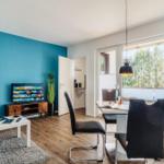 Wohn- und Essbereich mit TV, Tisch und Stühlen. Blaue Wand, Holzboden und Fenster mit Blick.