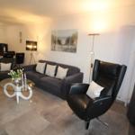 Living area with sofa, armchair, and glass table. Kitchen in background.