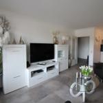 Living room with TV, glass table, and seating. Kitchen in the background.