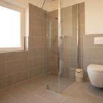 Badezimmer mit Dusche, WC und Fenster. Wand- und Bodenfliesen grau.