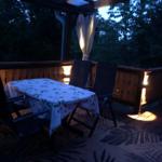 Terrasse mit Tisch, Stühlen und Beleuchtung. Hintergrund: dunkler Wald.