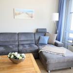 Ein grauer Sofa mit Kissen und Decke. Holztisch mit Blumen. Fenster mit Vorhängen.