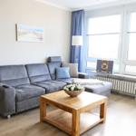 Ein Wohnzimmer mit grauem Sofa, Holztisch und Fenstern mit blauen Vorhängen.