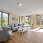 Wohnzimmer mit grauem Sofa, weißem Couchtisch und Holzboden. Große Fenster mit Blick auf Garten.