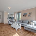 Wohnzimmer mit grauem Sofa, weißem Couchtisch und Holzboden. An der Wand hängen Strandbilder.