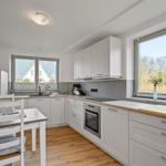 Küche mit weißen Schränken, Holzboden und Fenstern mit Blick auf Außenbereich.