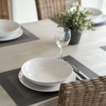 Tisch gedeckt mit weißen Tellern, Gläsern und dunklen Tischsets. Rattan-Stühle und Pflanze im Hintergrund.
