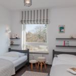 Two single beds with white linens and blankets. Window with striped roller blind. Wooden stools and wall shelves.