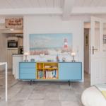 Wohnzimmer mit blauem Sideboard, Küchenbereich und Bett. Wandbild zeigt Leuchtturm.