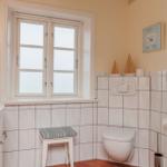 Badezimmer mit Toilette, Waschbecken und Fenster. Wandfliesen und Holzstuhl.