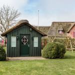 Ein grünes Holzhaus mit Strohdach und eine größere Villa mit Strohdach im Hintergrund.