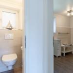 Badezimmer mit Toilette, Fenster und nautischem Dekor. Nebenraum mit Waschtisch und Heizkörper.