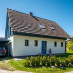 Ein Haus mit Schieferdach, blauer Tür und Garagenbereich. Grünflächen und Bäume im Hintergrund.