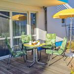 Terrasse mit Tisch, vier Stühlen und gelbem Sonnenschirm. Holzdielen und Pflanzen umgeben den Außenbereich.